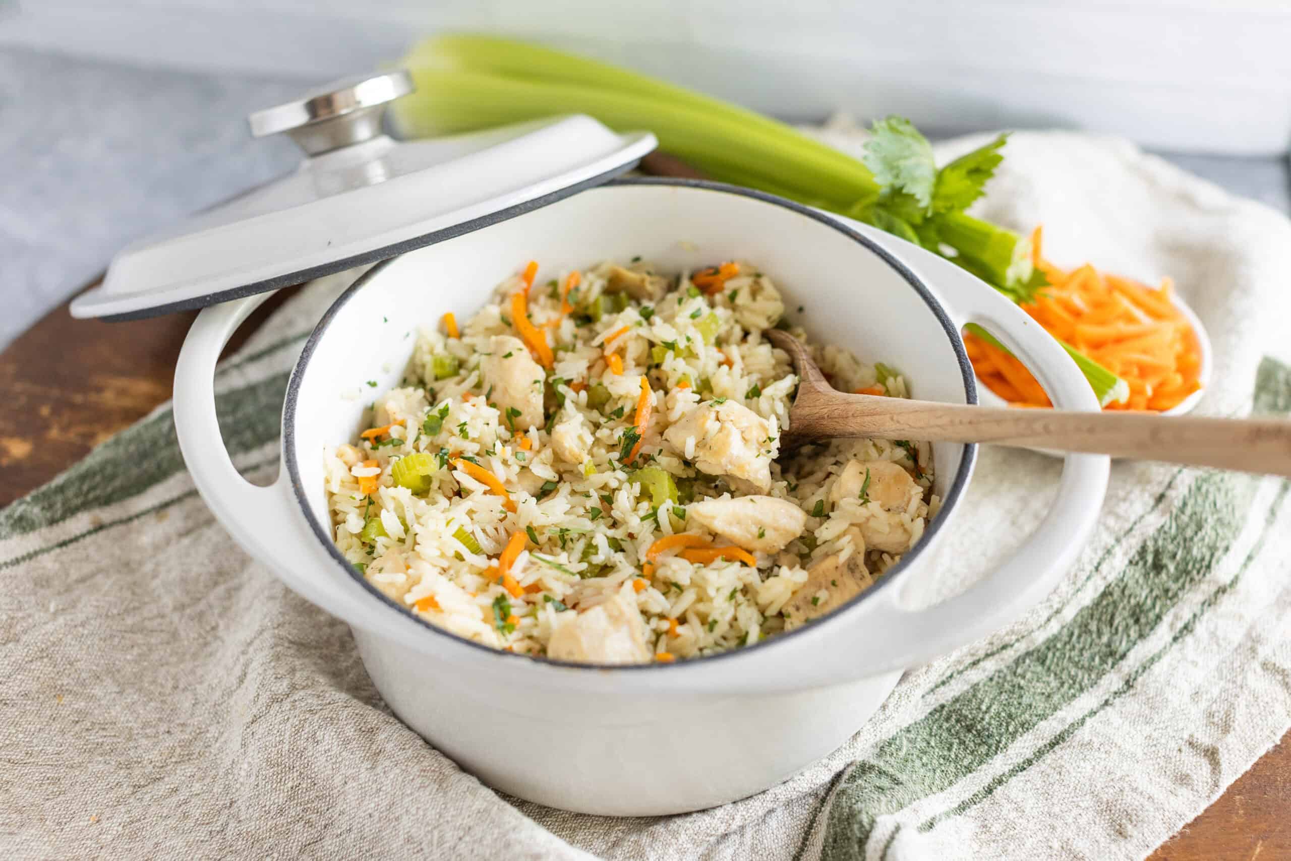 Steamed chicken rice in a white enamel pot with vegetables and herbs, on a rustic cloth with fresh celery and shredded carrots in the background.