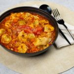 Savory tortellini soup with fresh vegetables in a rich tomato broth, served in a black bowl on a beige placemat with a fork and napkin for a delicious meal.