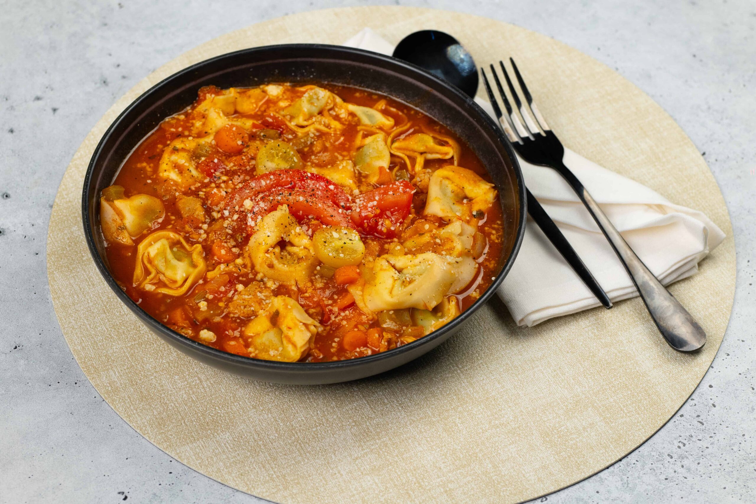 Savory tortellini soup with fresh vegetables in a rich tomato broth, served in a black bowl on a beige placemat with a fork and napkin for a delicious meal.