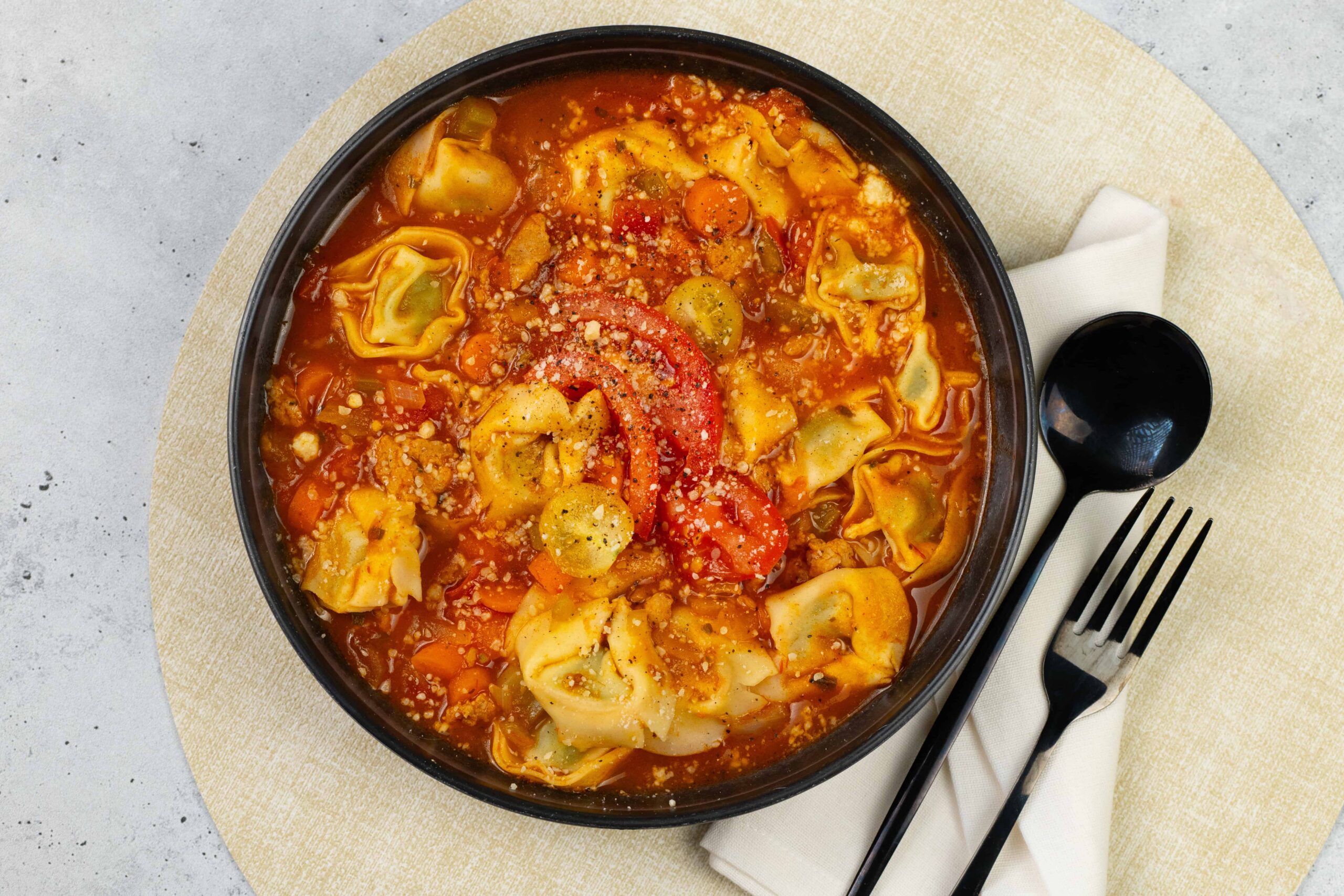 Hearty homemade tortellini soup in a rich tomato broth with vegetables and grated cheese, served in a black bowl, perfect for comforting meals and easy weeknight dinner ideas.