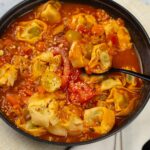 Savory tortellini soup in a black bowl with tomato broth, vegetables, and grated cheese on top, served with a fork and cloth napkin. Perfect comfort food for cold days.