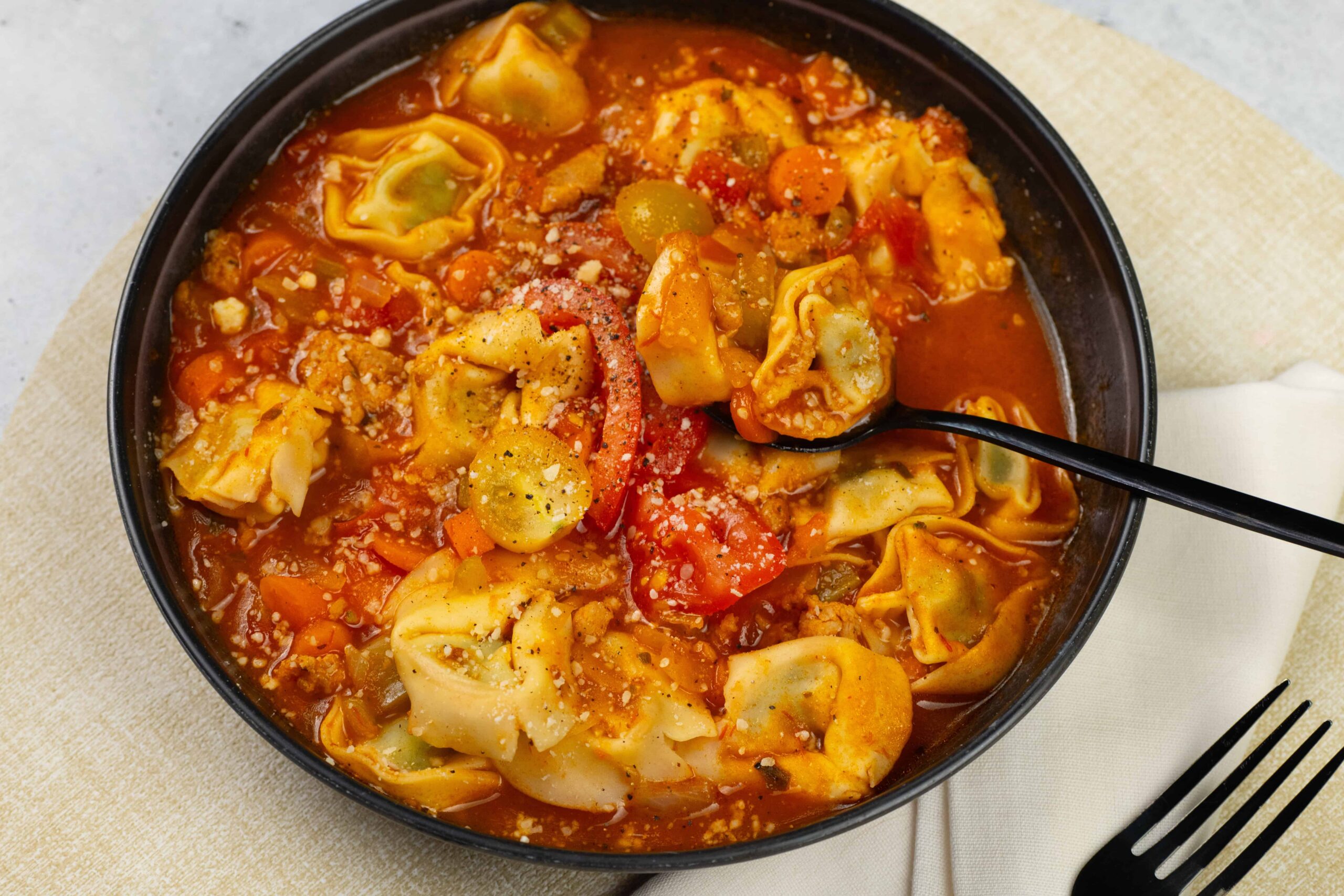 Savory tortellini soup in a black bowl with tomato broth, vegetables, and grated cheese on top, served with a fork and cloth napkin. Perfect comfort food for cold days.