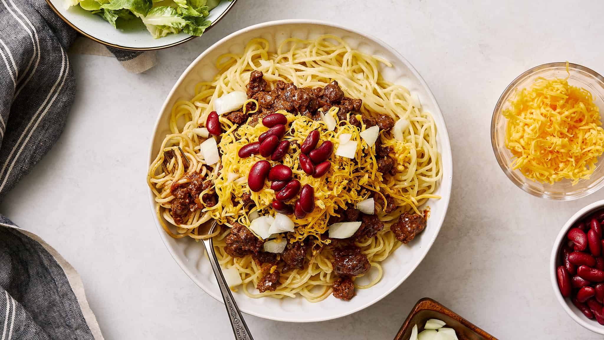 Savory beef spaghetti with kidney beans and shredded cheese on a white plate, topped with chopped onions, served with side bowls of shredded cheese and kidney beans, perfect for easy dinner recipes and comfort food.