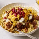 Cheesy beef chili spaghetti bake topped with kidney beans and shredded cheese, served in a white bowl with fresh onion and lettuce on the side, perfect for a hearty meal.