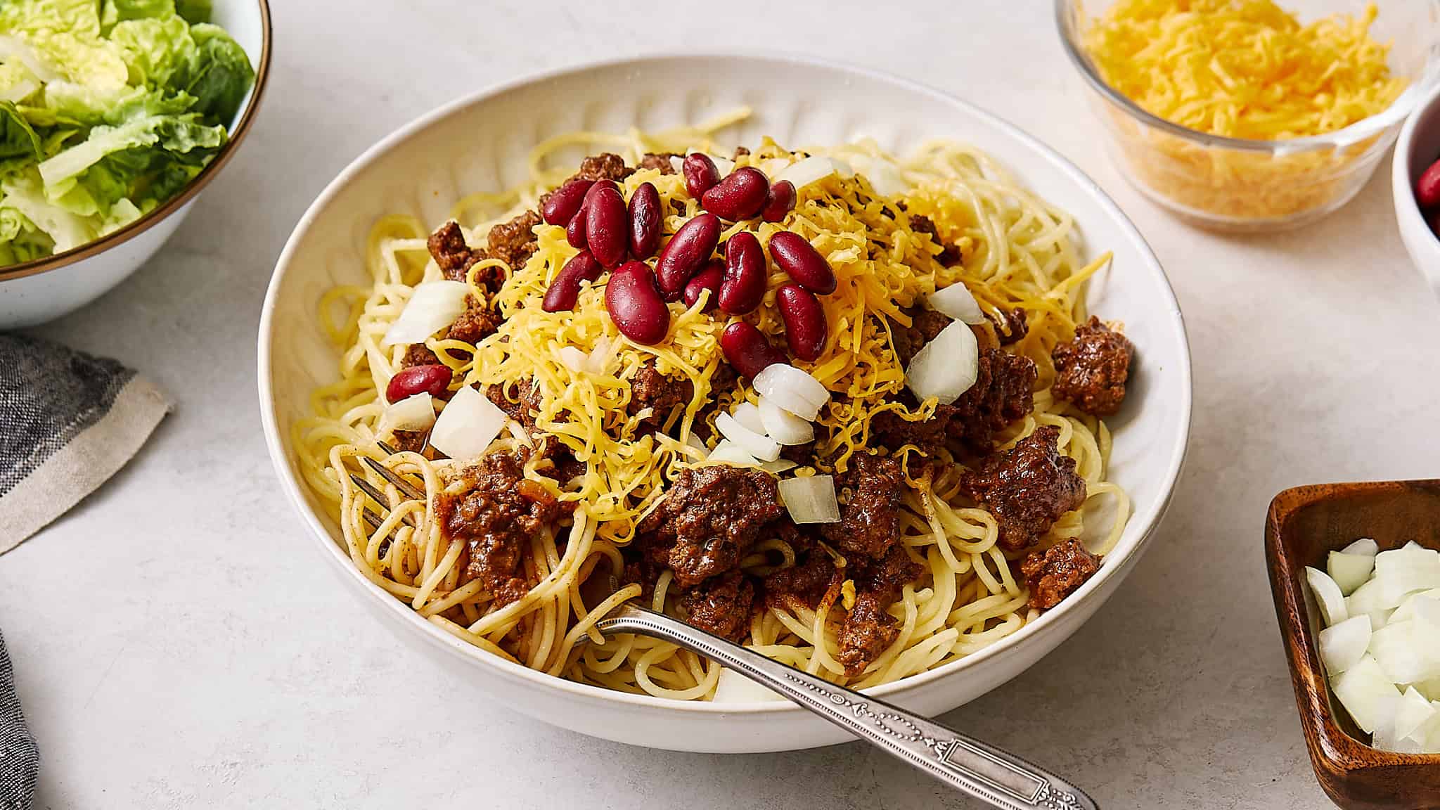 Cheesy beef chili spaghetti bake topped with kidney beans and shredded cheese, served in a white bowl with fresh onion and lettuce on the side, perfect for a hearty meal.