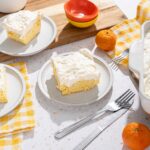 Fluffy lemon cake with creamy frosting on white plates, served on a speckled countertop with fresh oranges and colorful bowls, perfect for spring desserts.