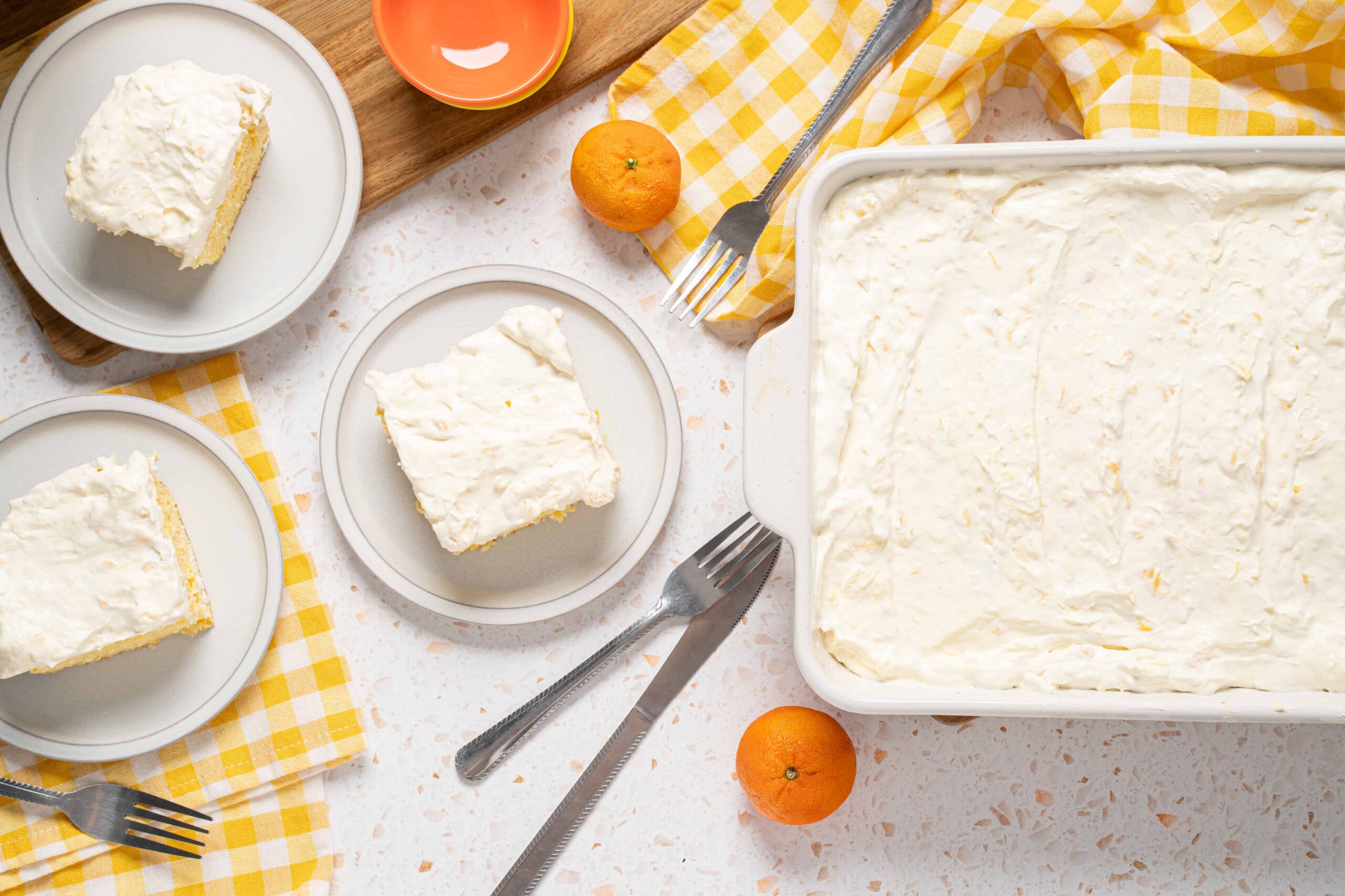 Creamy lemon cake slices with fluffy frosting served on white plates, fresh oranges on a yellow checkered cloth, and a baking dish of leftover cake, perfect for dessert recipes and citrus-flavored treats.