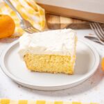 Fluffy lemon cake slice with creamy frosting on a white plate, surrounded by fresh oranges, a yellow checkered cloth, and cutlery for a delicious dessert presentation.