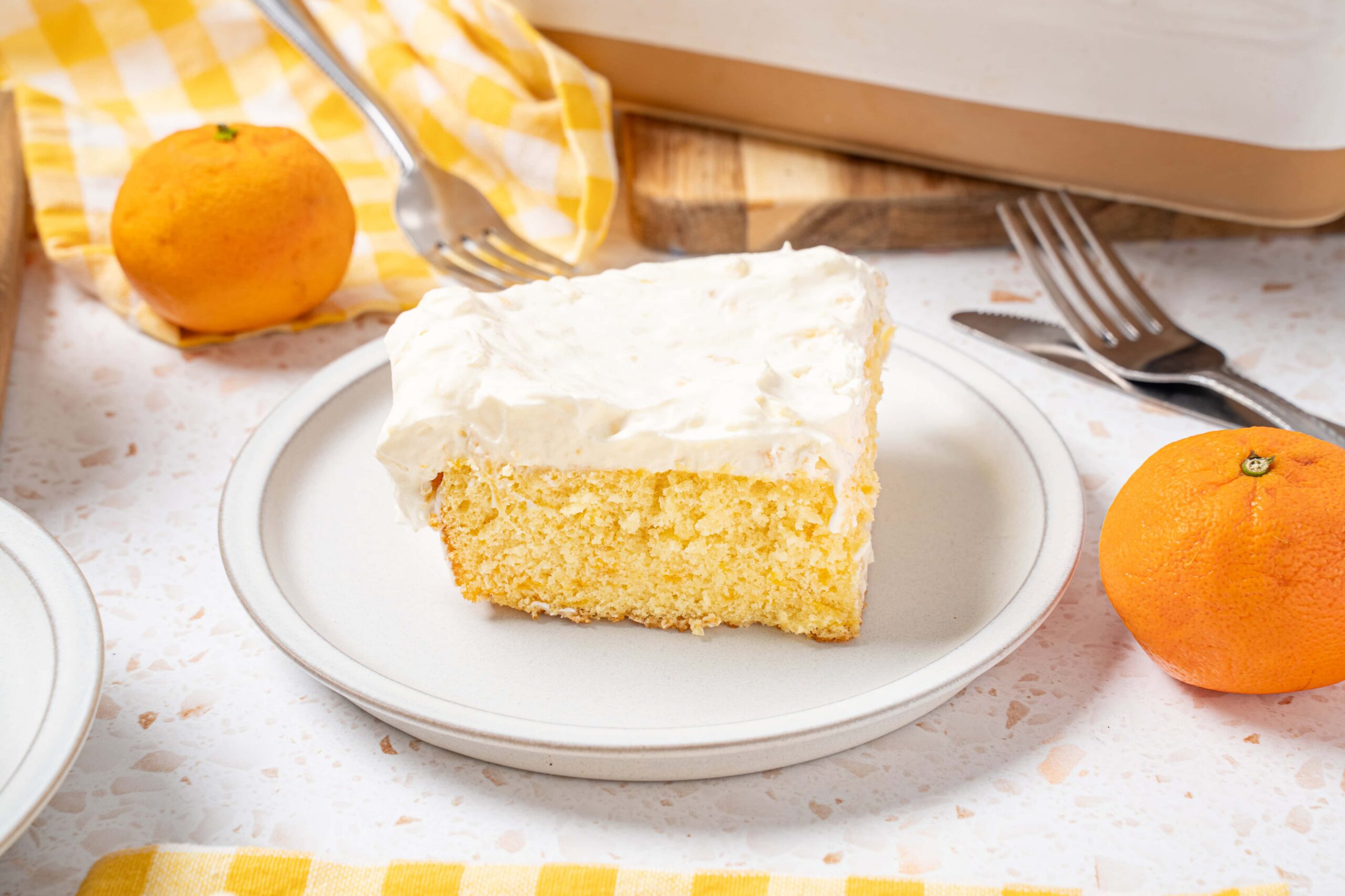 Fluffy lemon cake slice with creamy frosting on a white plate, surrounded by fresh oranges, a yellow checkered cloth, and cutlery for a delicious dessert presentation.