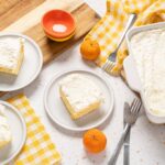 Fluffy lemon cake slices with creamy frosting on white plates, fresh mandarins, and baking ingredients on a white countertop with yellow checkered napkins.