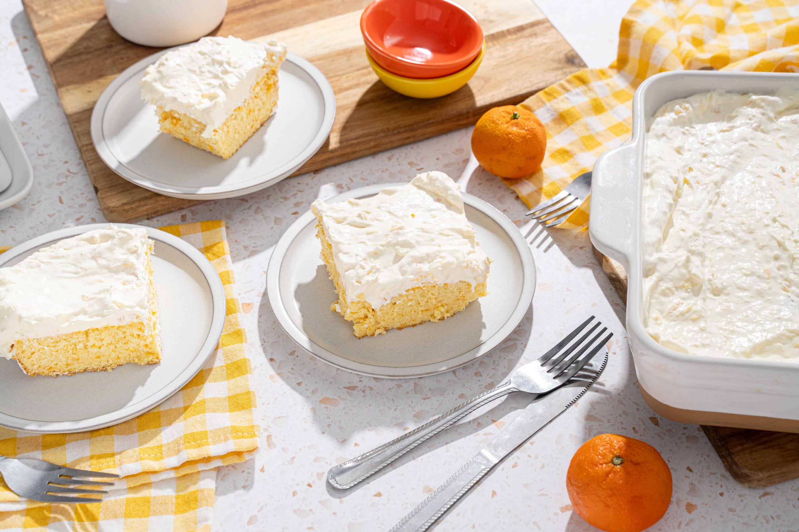 Fluffy lemon cake with creamy frosting on white plates, served on a speckled countertop with fresh oranges and colorful bowls, perfect for spring desserts.
