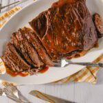 Juicy glazed beef brisket on a white serving platter with a fork, served on a checkered cloth, perfect for roast beef recipes or holiday dinners.