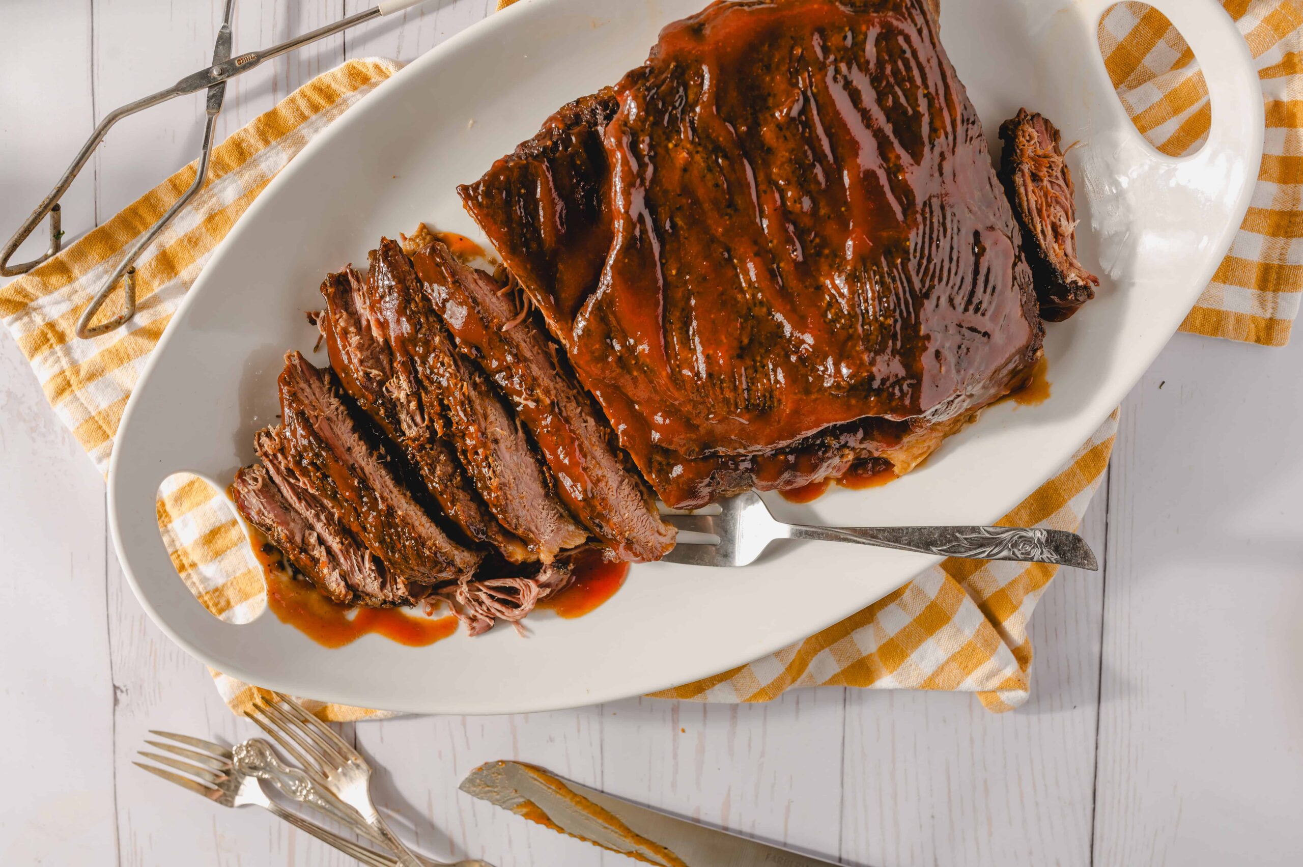 Juicy glazed beef brisket on a white serving platter with a fork, served on a checkered cloth, perfect for roast beef recipes or holiday dinners.