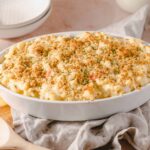 Creamy macaroni and cheese casserole topped with breadcrumbs, served in a white oval dish on a rustic wooden board with a linen napkin background.