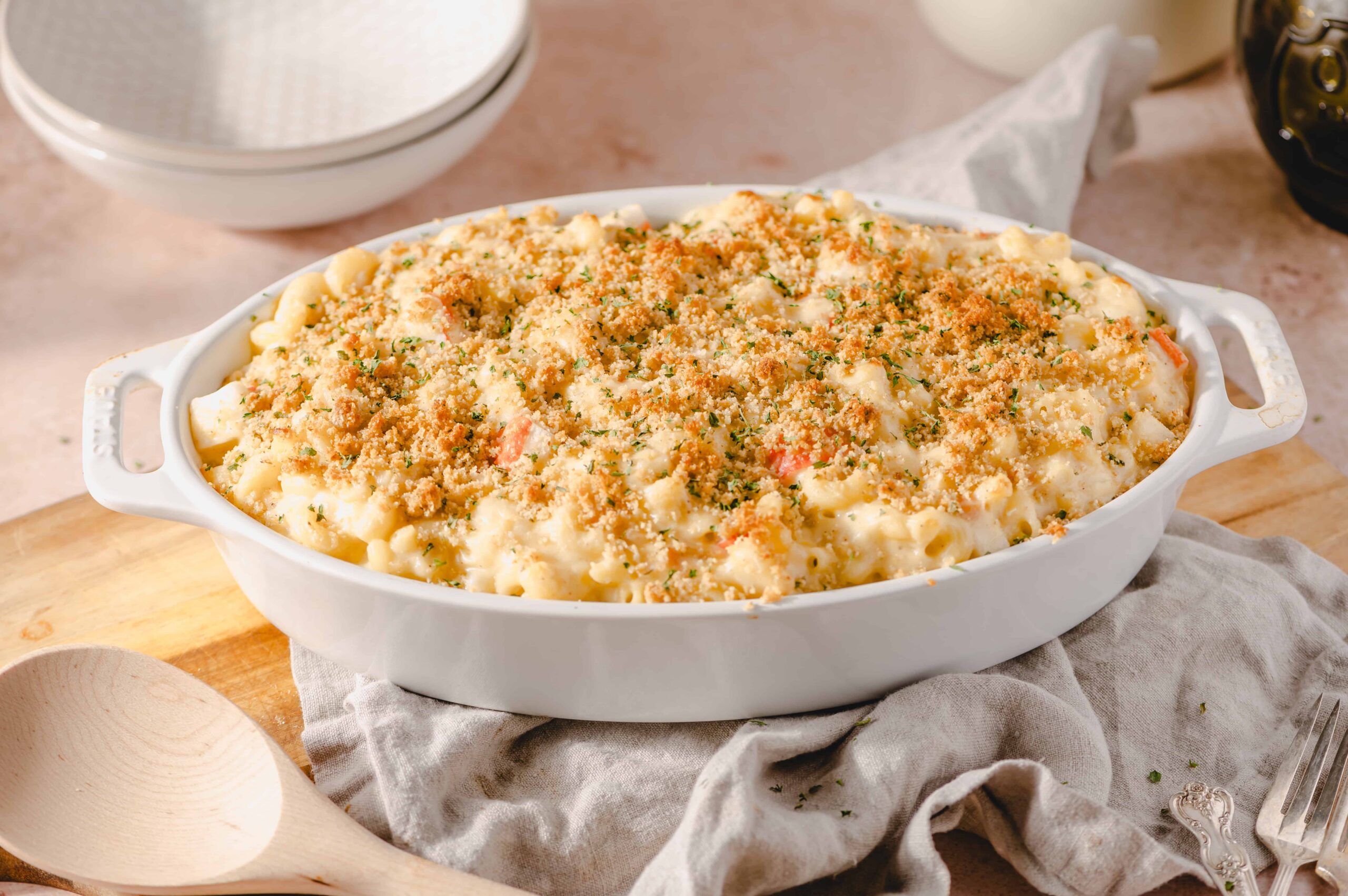 Creamy macaroni and cheese casserole topped with breadcrumbs, served in a white oval dish on a rustic wooden board with a linen napkin background.