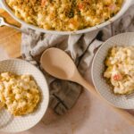 Creamy cauliflower casserole with breadcrumb topping, served in white patterned bowls on a textured pink surface. Includes a large baking dish, wooden spoon, and vintage forks for a rustic presentation.