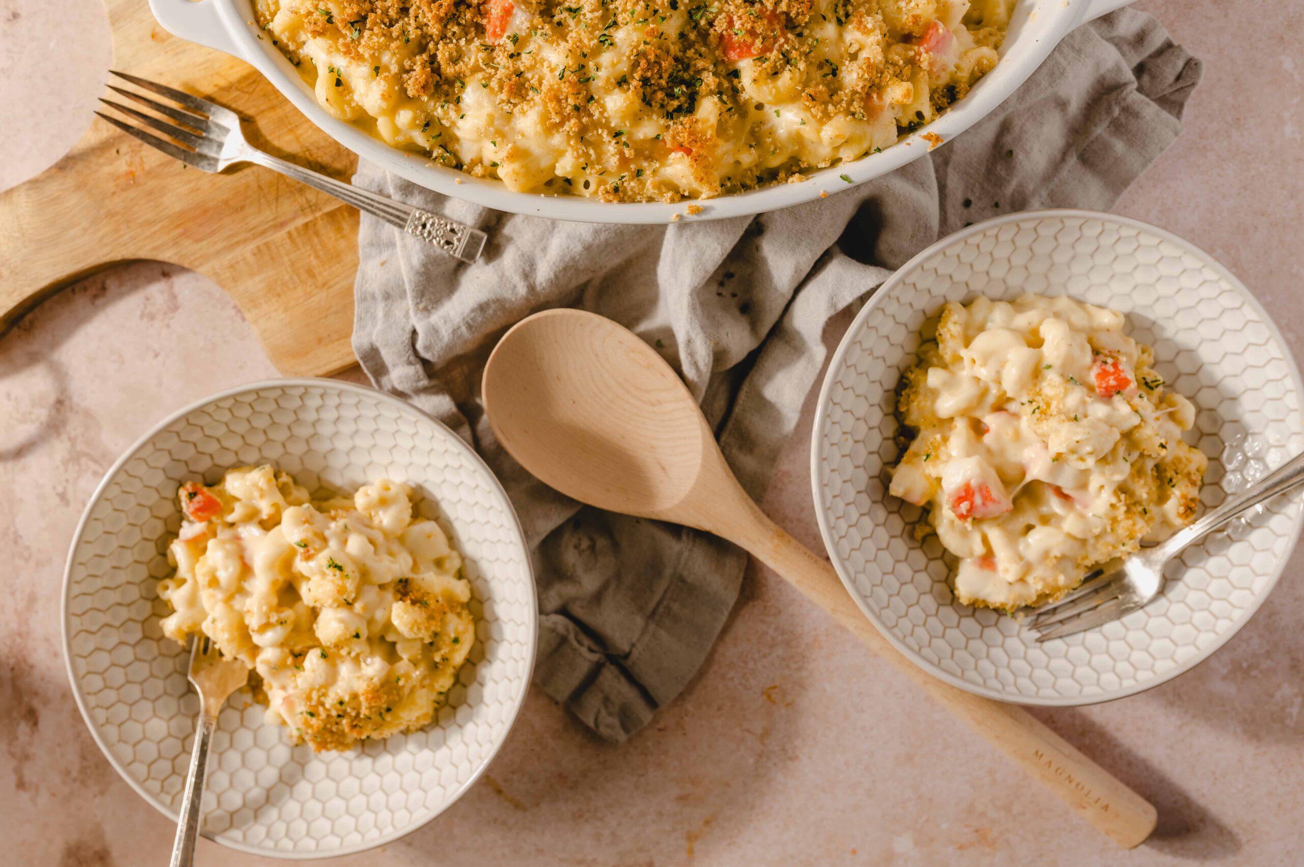 Creamy cauliflower casserole with breadcrumb topping, served in white patterned bowls on a textured pink surface. Includes a large baking dish, wooden spoon, and vintage forks for a rustic presentation.