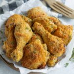 Crispy baked chicken drumsticks with golden breadcrumb coating, served on a piece of parchment paper with fresh thyme herbs. Perfect for a family dinner or casual outdoor meal.