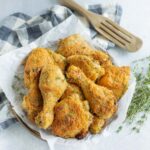 Crispy baked chicken drumsticks with golden breadcrumb coating on a white crumpled parchment paper, garnished with fresh thyme, served on a rustic plate with a checkered napkin and wooden serving fork for a delicious homemade meal.