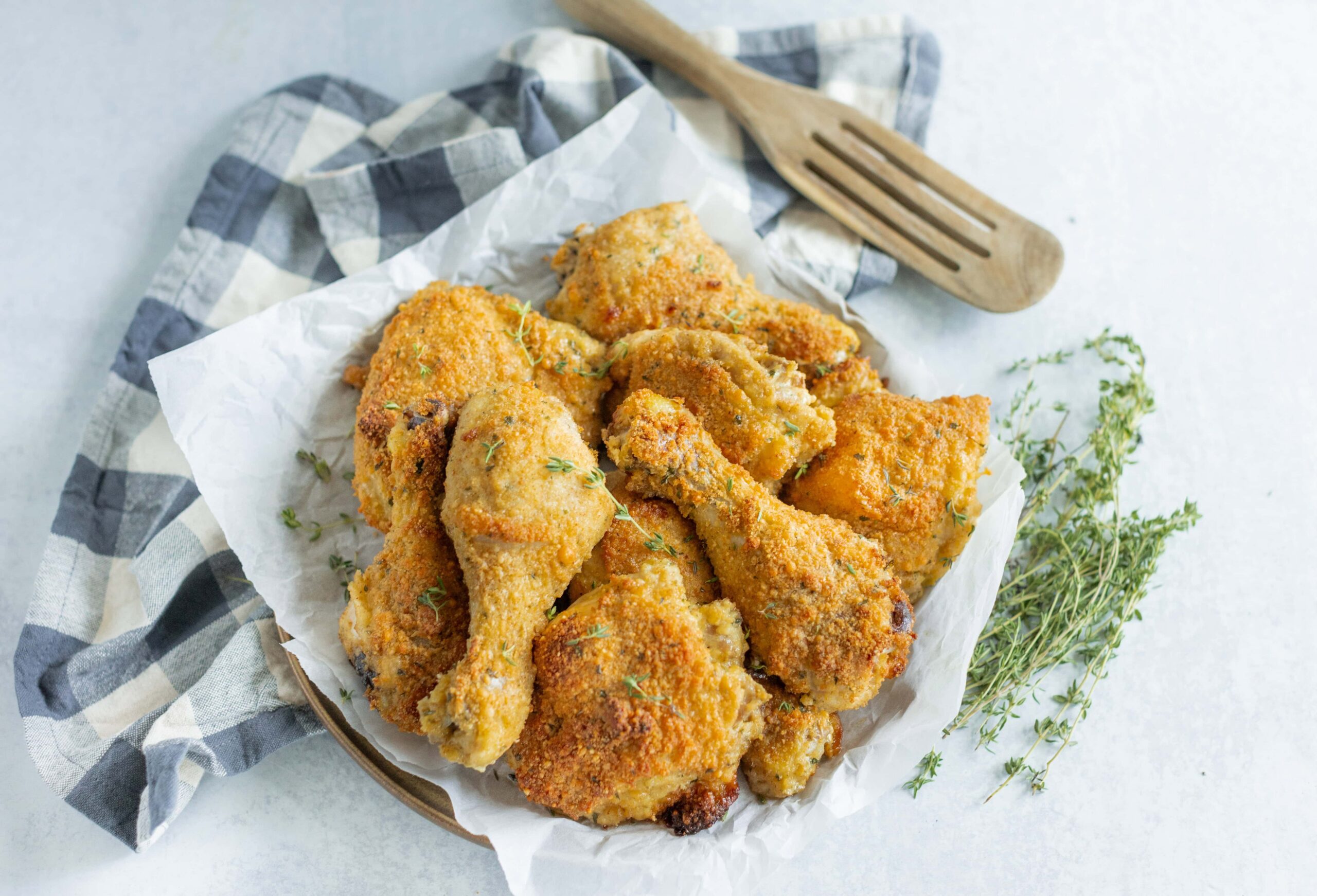 Crispy baked chicken drumsticks with golden breadcrumb coating on a white crumpled parchment paper, garnished with fresh thyme, served on a rustic plate with a checkered napkin and wooden serving fork for a delicious homemade meal.