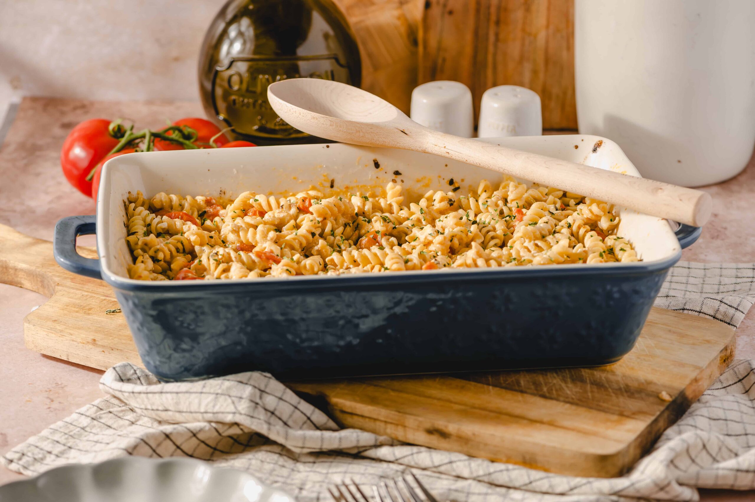 Creamy baked pasta casserole with rotini, cherry tomatoes, and herbs in a blue ceramic dish on a wooden cutting board, ready to serve.