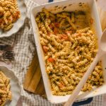 Creamy baked rotini pasta with tomatoes and herbs, served in a casserole dish. Fresh tomatoes and pasta leftovers on plates and a wooden table setting. Perfect comfort food recipe.