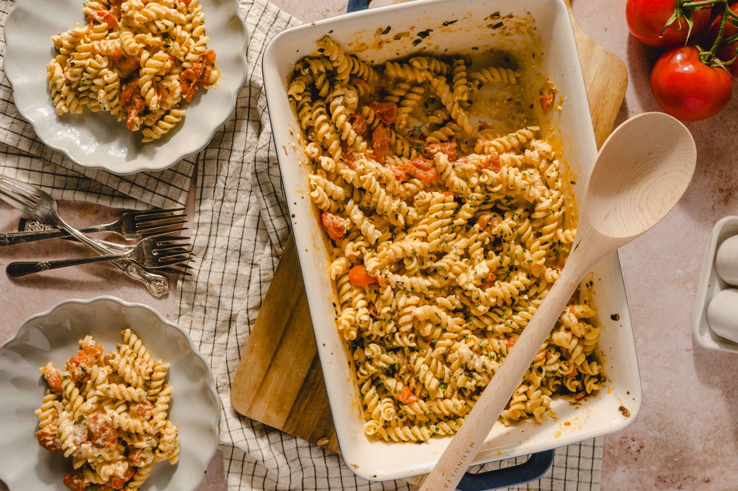 Creamy baked rotini pasta with tomatoes and herbs, served in a casserole dish. Fresh tomatoes and pasta leftovers on plates and a wooden table setting. Perfect comfort food recipe.