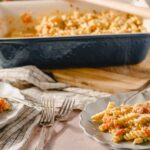 Creamy pasta with cherry tomatoes and herbs served on white plates, with a baked pasta casserole dish in the background on a wooden surface for comfort food recipes.