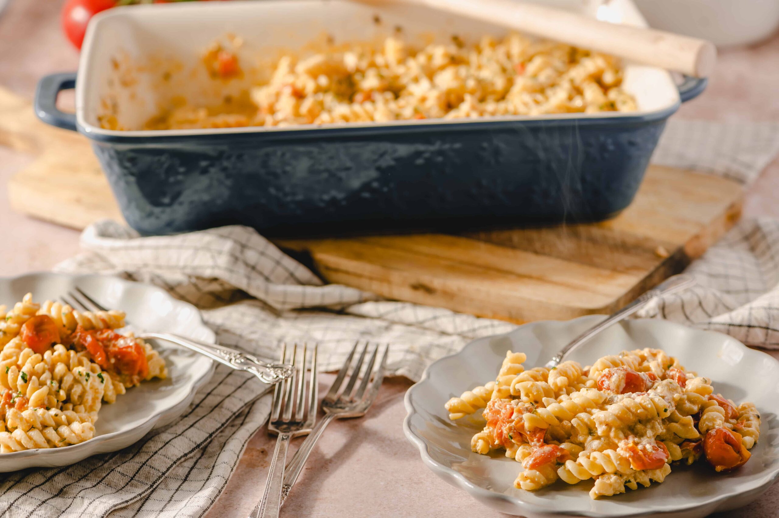 Creamy pasta with cherry tomatoes and herbs served on white plates, with a baked pasta casserole dish in the background on a wooden surface for comfort food recipes.