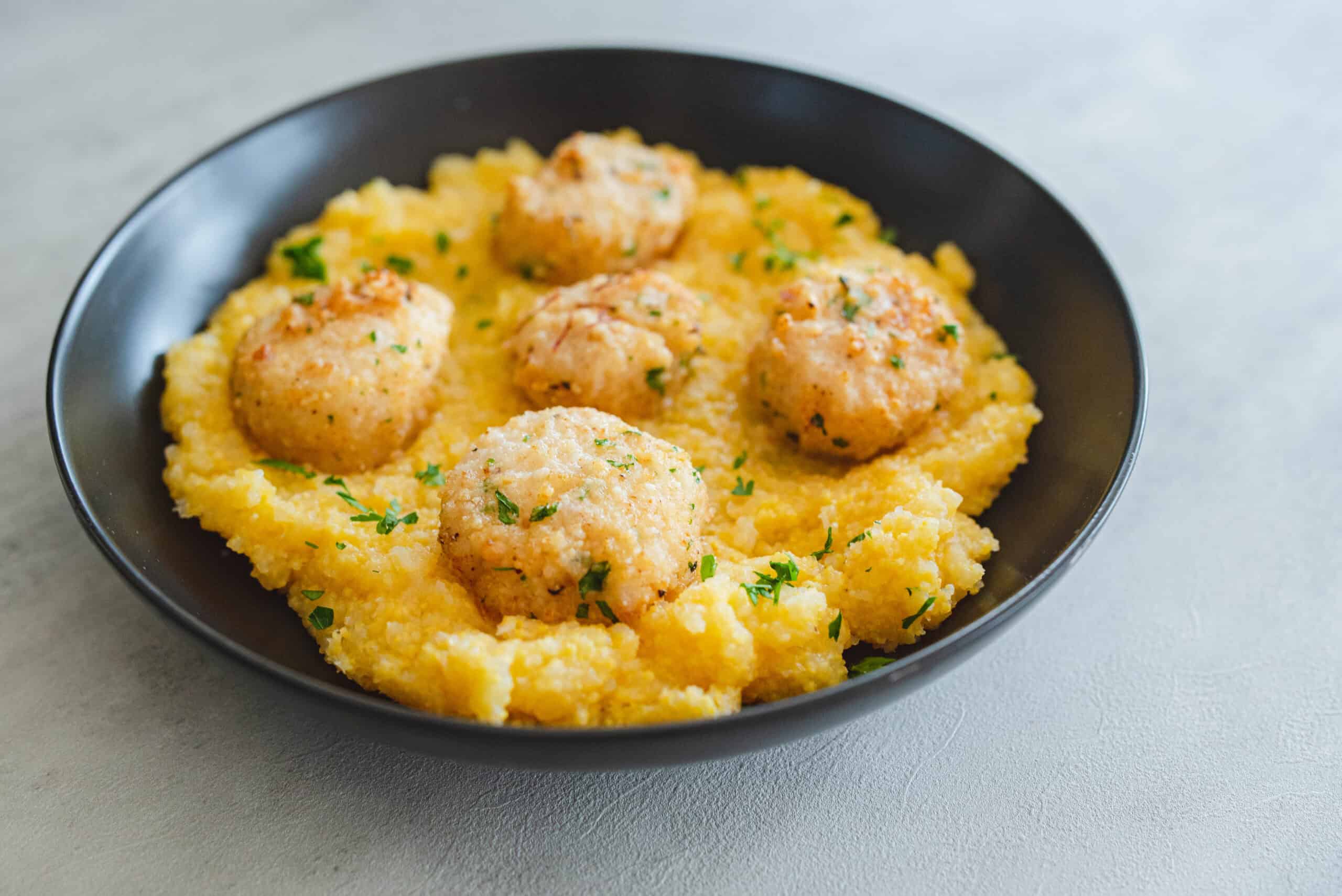 Savory chicken and herb meatballs served over creamy polenta garnished with fresh parsley in a black bowl. Perfect comfort food recipe for family dinner or special occasion.