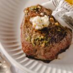 Juicy grilled steak with melted butter and herbs served on white plate, with a side of crispy roasted potatoes in parchment paper, perfect for steak recipe and gourmet dinner ideas.