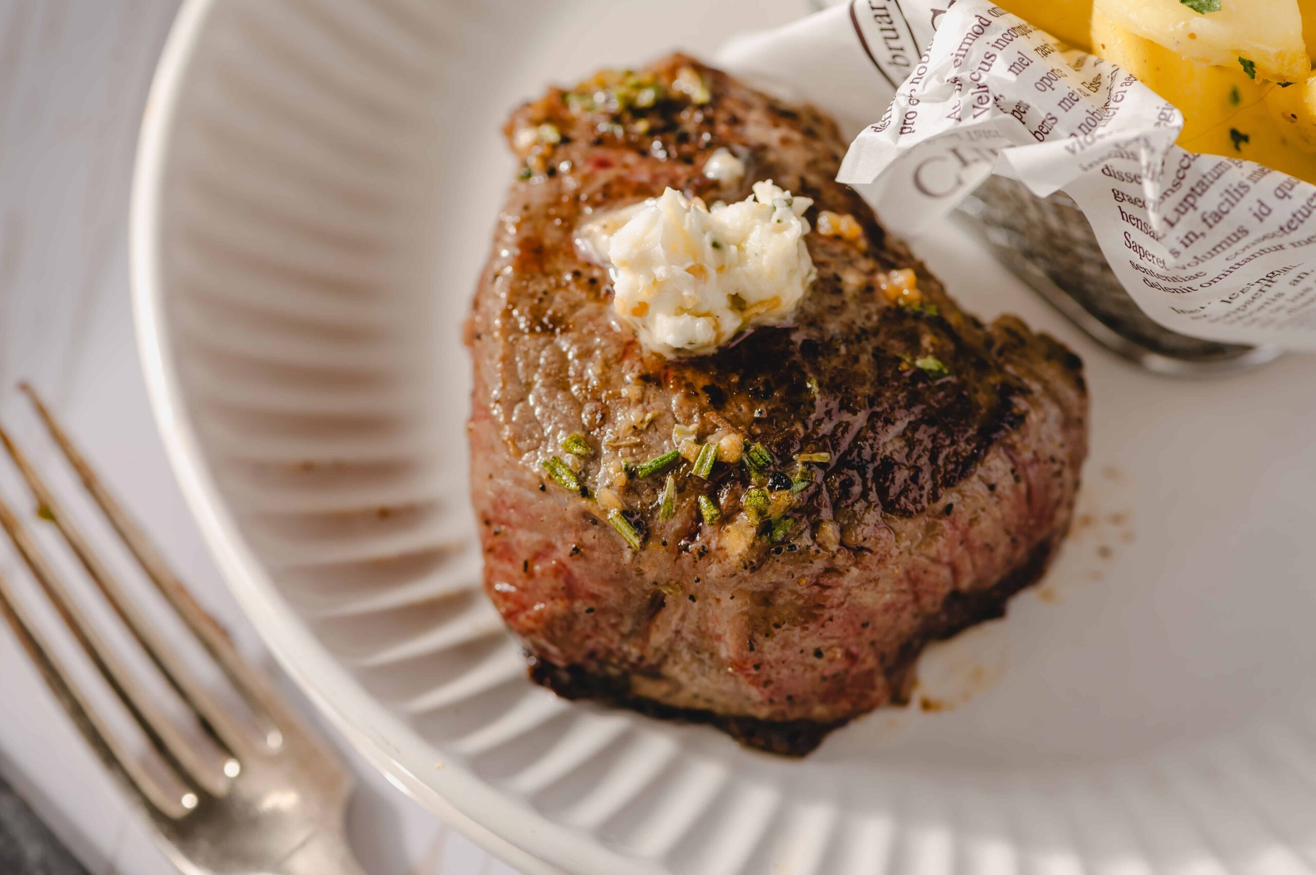 Juicy grilled steak with melted butter and herbs served on white plate, with a side of crispy roasted potatoes in parchment paper, perfect for steak recipe and gourmet dinner ideas.