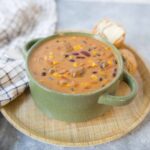 Creamy bean and corn chili served in a green speckled mug on a wooden tray with bread rolls and butter on the side, perfect for a hearty, comforting meal.