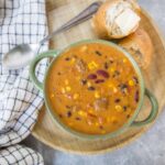 Creamy beef and bean chili with corn and kidney beans in a green speckled bowl served with bread on a wooden platter.
