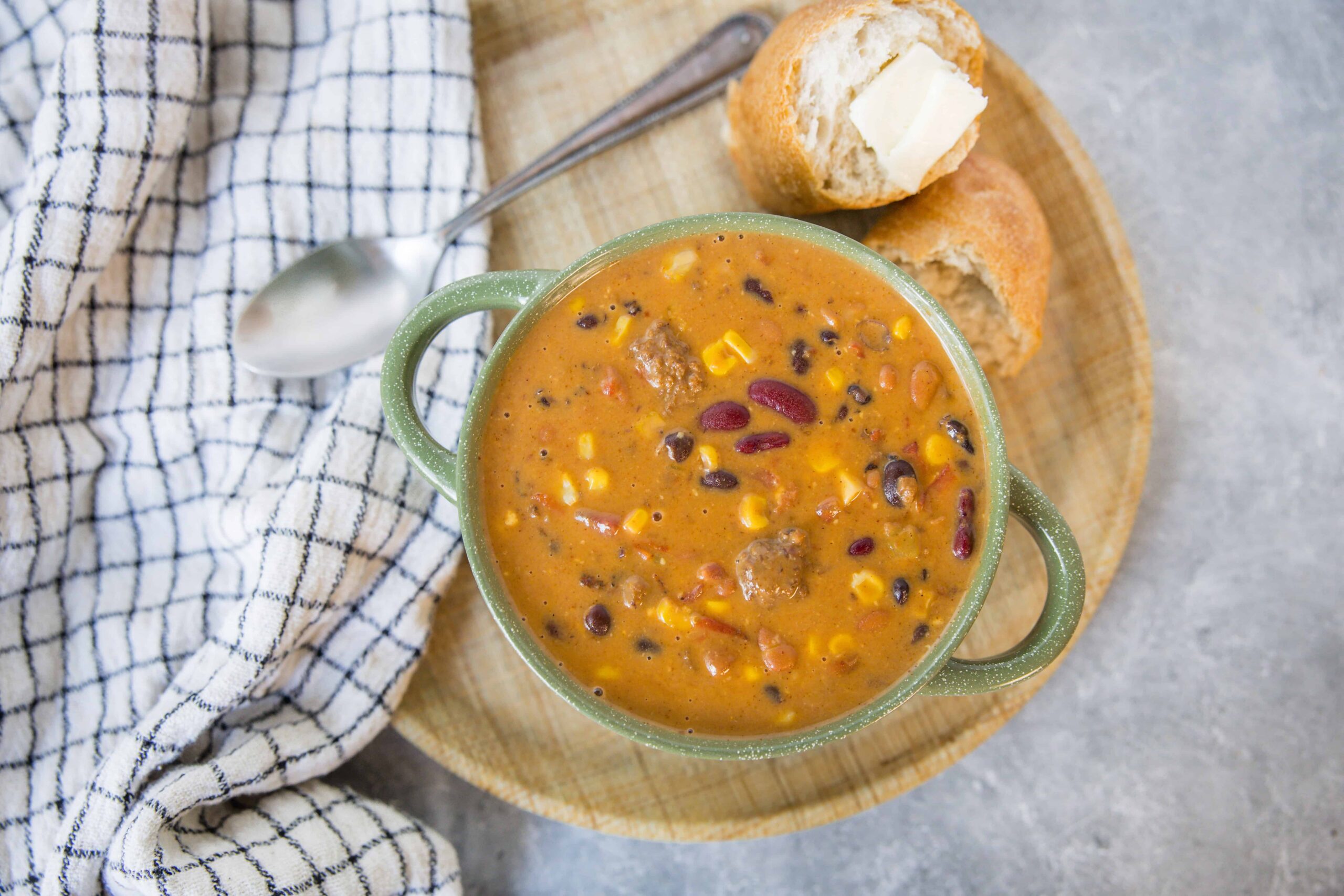Creamy beef and bean chili with corn and kidney beans in a green speckled bowl served with bread on a wooden platter.