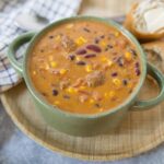 Creamy vegetable chili soup with beans and corn served in a rustic green mug, accompanied by bread rolls with butter on a wooden tray, perfect for hearty comfort food; popular recipe on Hey Recipe.