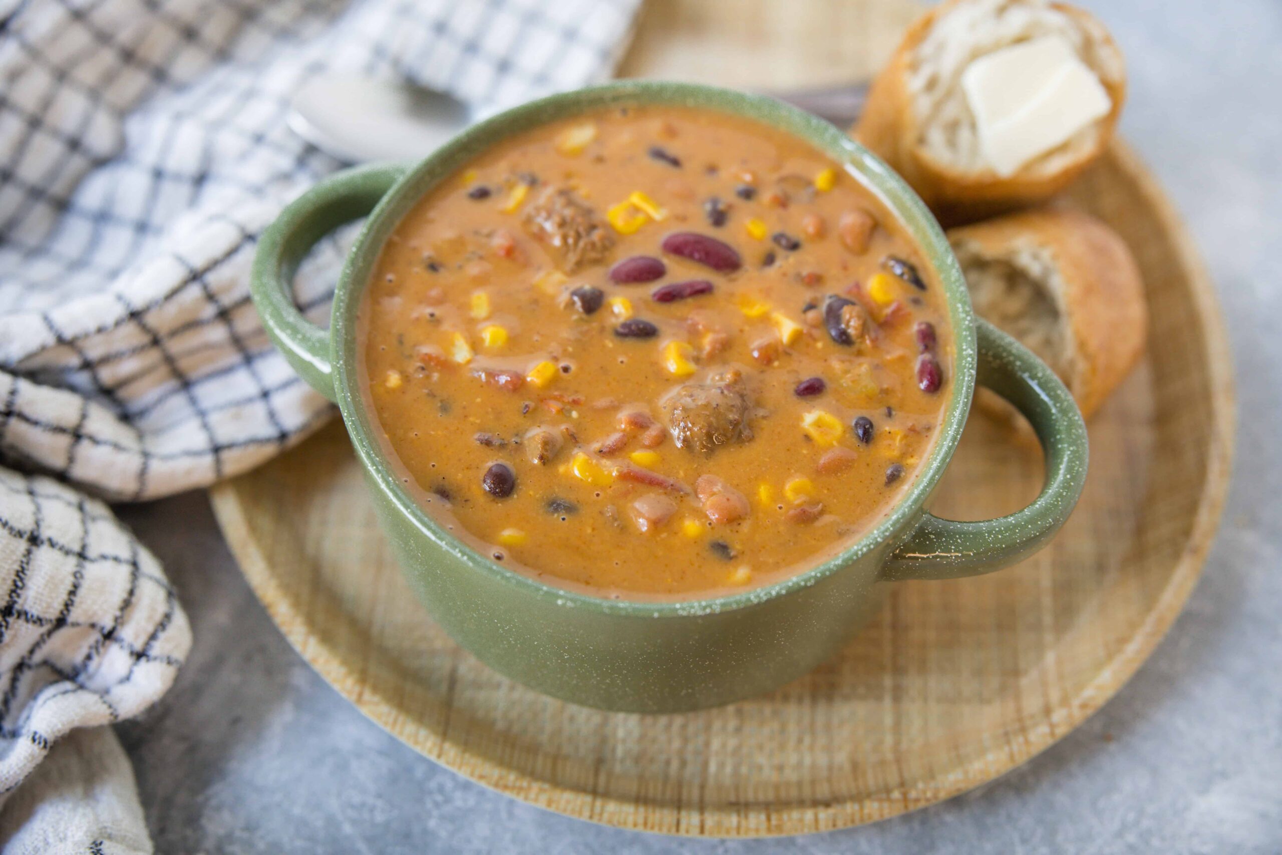 Creamy vegetable chili soup with beans and corn served in a rustic green mug, accompanied by bread rolls with butter on a wooden tray, perfect for hearty comfort food; popular recipe on Hey Recipe.