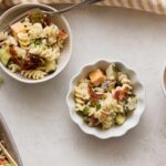 Creamy pasta salad with sweet potatoes, bacon, cucumbers, and green onions in small white bowls on a light textured surface.