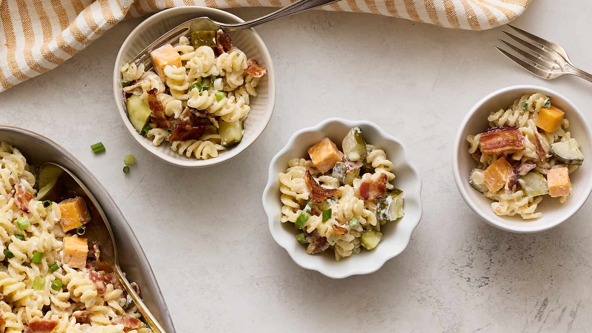 Creamy pasta salad with sweet potatoes, bacon, cucumbers, and green onions in small white bowls on a light textured surface.