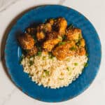Sweet and spicy orange chicken served over fluffy white rice garnished with chopped green onions on a rustic blue plate.