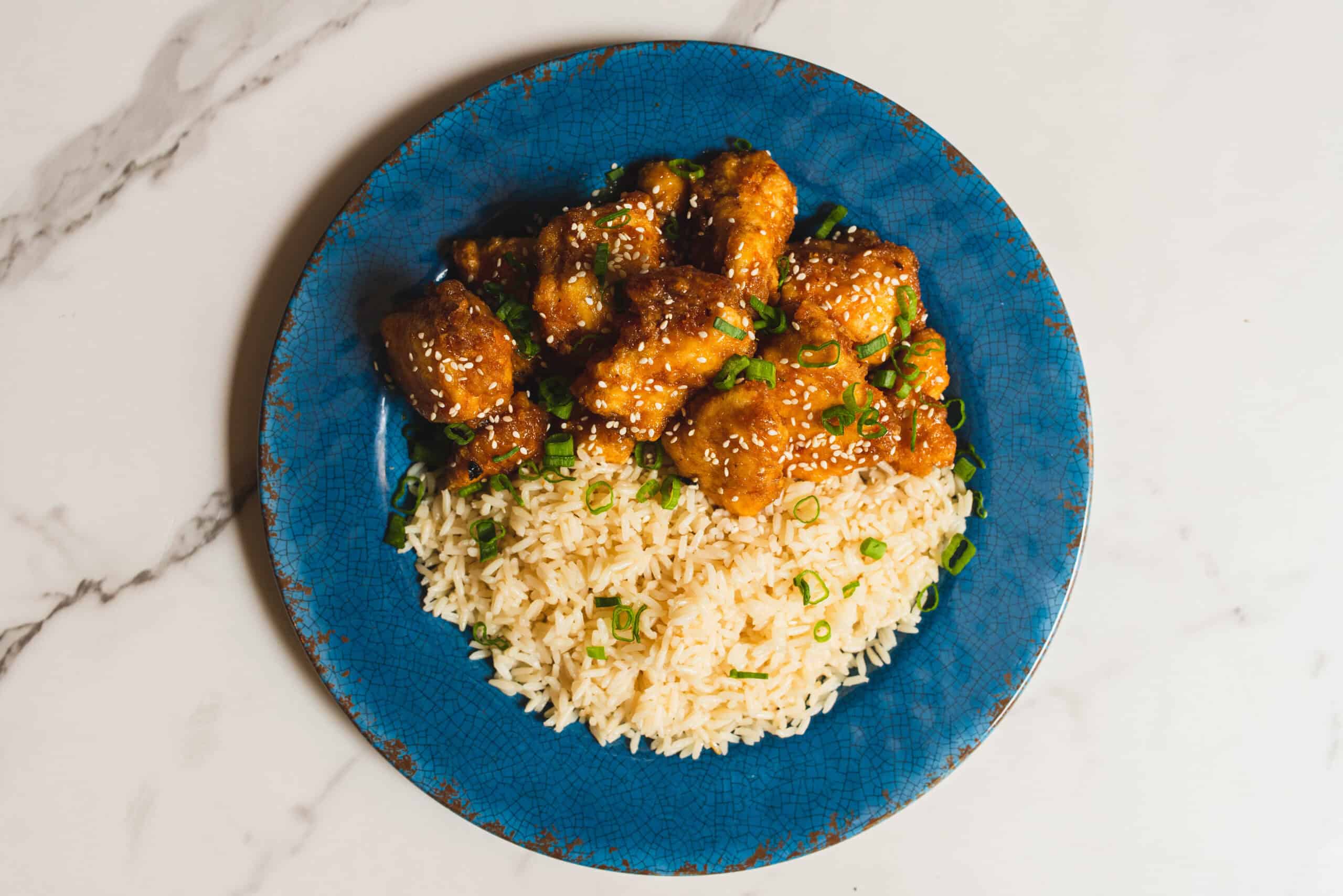 Sweet and spicy orange chicken served over fluffy white rice garnished with chopped green onions on a rustic blue plate.