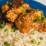 Crispy fried chicken pieces coated in orange sauce, garnished with sesame seeds and chopped green onions, served over white rice on a blue plate.