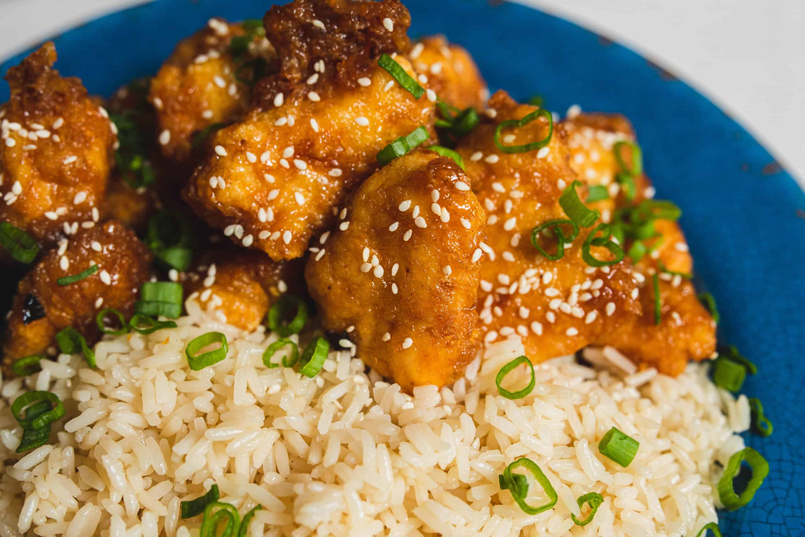 Crispy fried chicken pieces coated in orange sauce, garnished with sesame seeds and chopped green onions, served over white rice on a blue plate.
