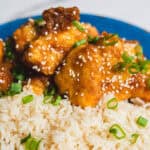Crispy fried chicken bites glazed with sauce, garnished with sesame seeds and chopped green onions, served over fluffy white rice on a vibrant blue plate.