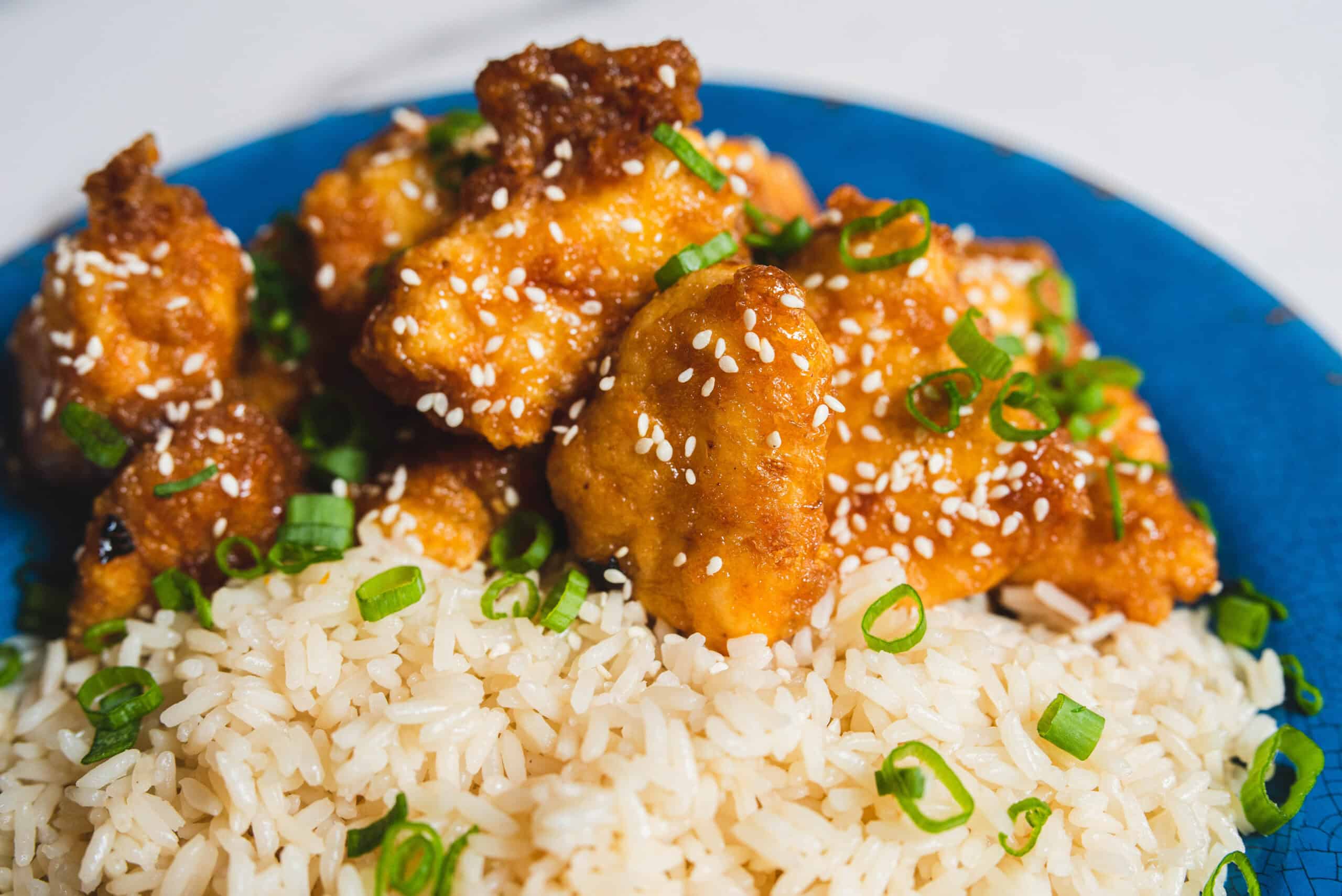 Crispy fried chicken bites glazed with sauce, garnished with sesame seeds and chopped green onions, served over fluffy white rice on a vibrant blue plate.