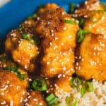 Crispy buffalo chicken bites garnished with sesame seeds and chopped green onions served with steamed rice in a blue bowl.
