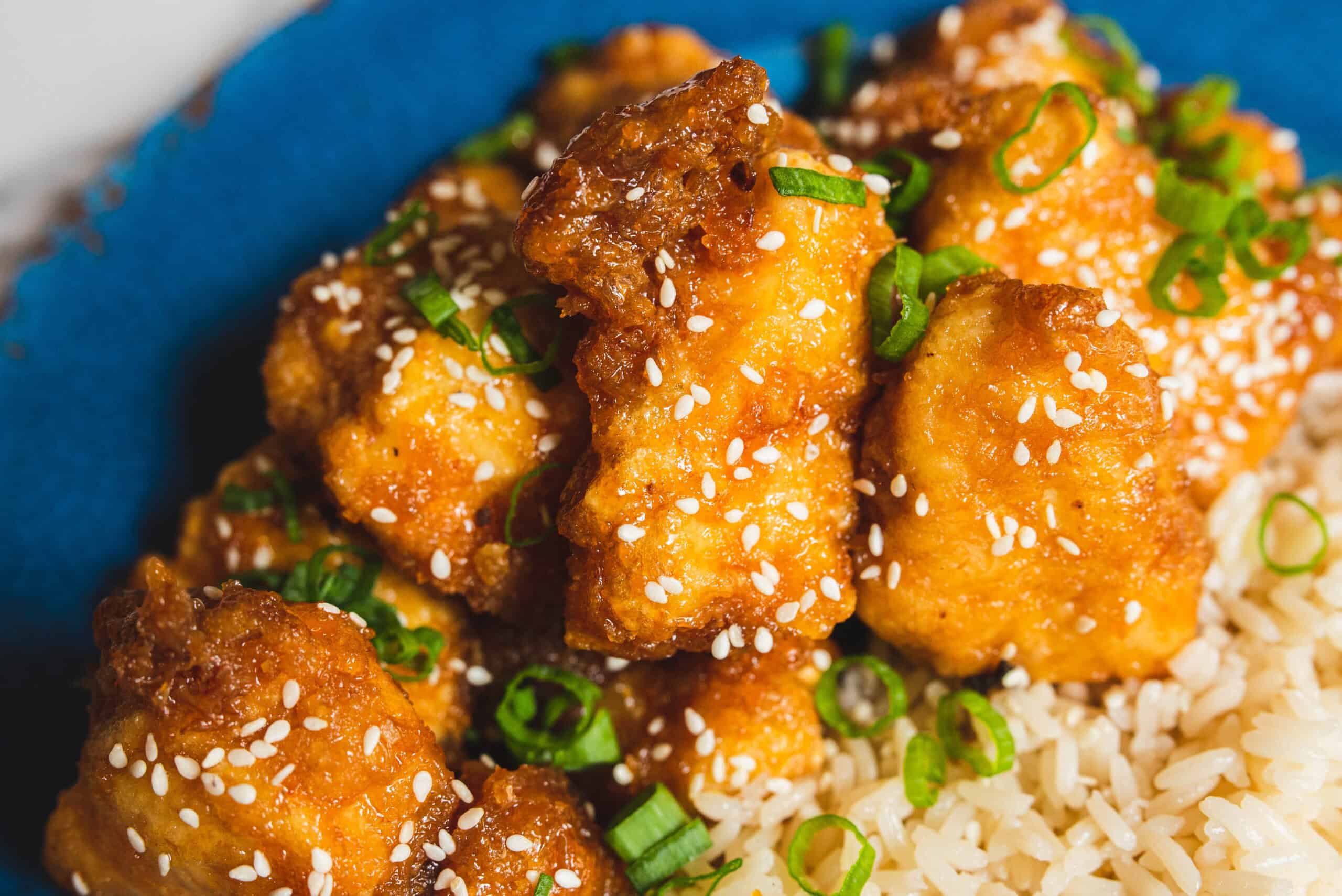 Crispy buffalo chicken bites garnished with sesame seeds and chopped green onions served with steamed rice in a blue bowl.