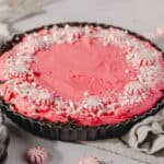Pink peppermint pie with whipped cream and crushed peppermint candies in a tart pan on a white surface.