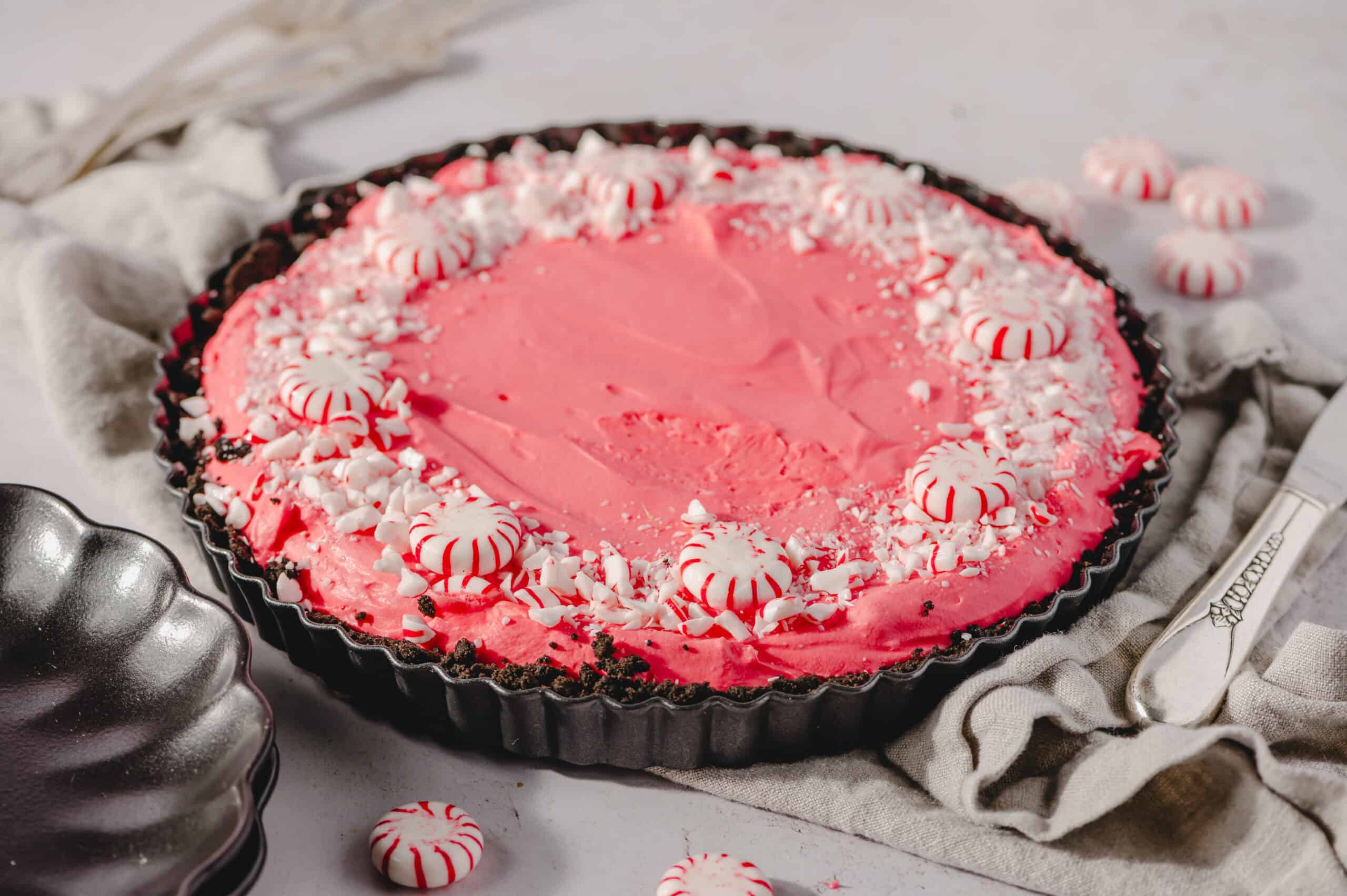 Pink peppermint pie with whipped cream and crushed peppermint candies in a tart pan on a white surface.