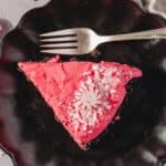 Delicious peppermint chocolate cheesecake slice with pink frosting, peppermint candy, and white chocolate shavings on a black decorative plate. Perfect for holiday desserts and festive occasions.